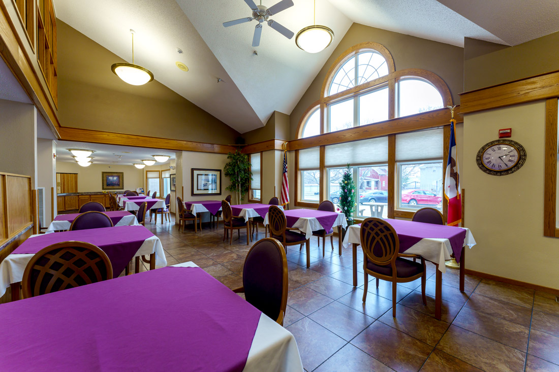 Ostego place dining area with square tables with purple and white tablecloths, and large windows.
